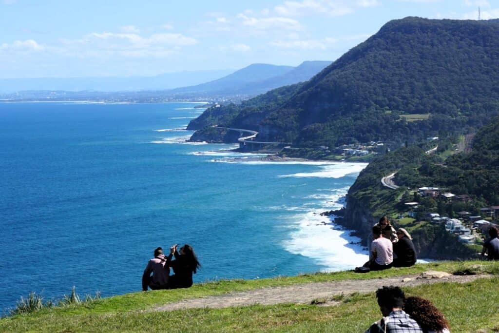 People sitting on the grass enjoying Stunning coastal views of cliffs and blue ocean and surf at Bald Hill Lookout