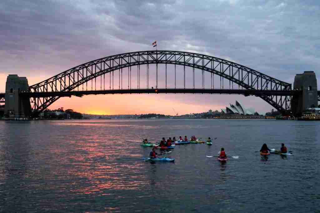 The Sydney Harbour bridge and opera house with the sunrising behind the harbour bridge and kayaks on the water