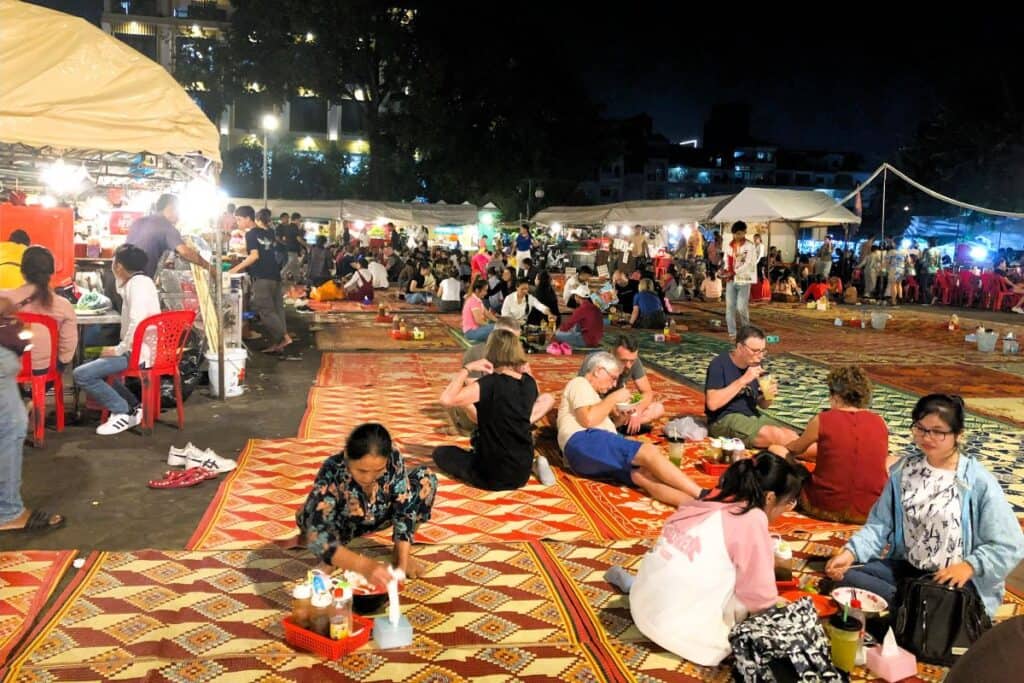 People eating on colourful mats on the groundsurrounded by market stalls at the Phnom Penh Night Market