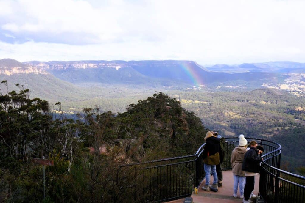 A group of people standing at Hargraves lookout with views of forwsted valleys and escarpment and a rainbow