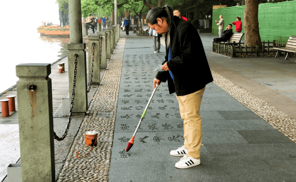 a man with a large brush painting with water on the pavement beside a lake 